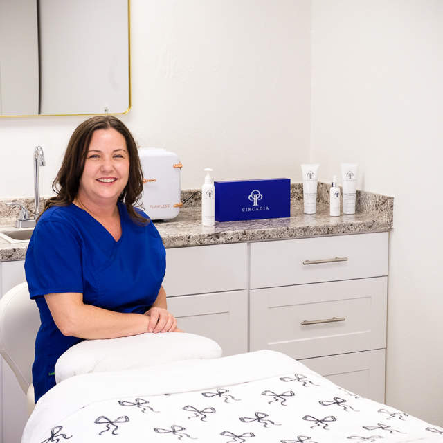 Sonya Dilbeck, an esthetician, is seated on a stool
							behind her treatment table. Behind her is a counter that displays some of her treatment
							products and a blue Circadia promotional plaque.